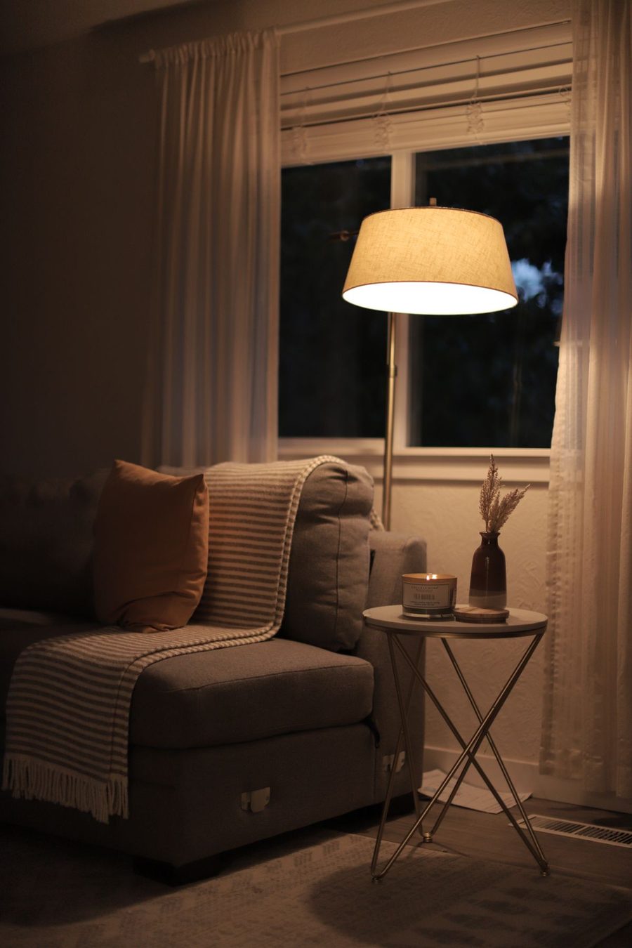 A living room at dusk: a warm floor lamp and a candle glow softly while the big overhead light stays off